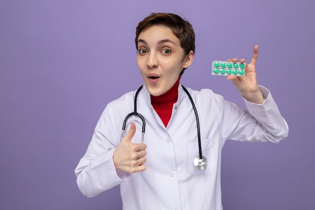 Happy and surprised young woman doctor in white coat with stethoscope holding blister with pills looking smiling cheerfully showing thumbs up
