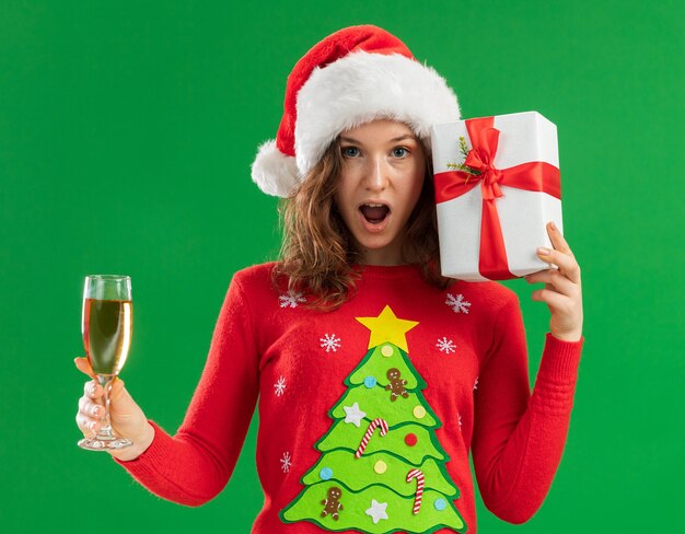 Happy and surprised young woman in red christmas sweater  and santa hat holding glass of champagne and present looking at camera cheerfully standing over green  background