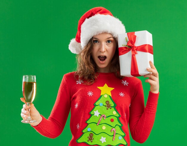 Счастливая и удивленная молодая женщина в красном рождественском свитере и шляпе санта-клауса, держащая бокал шампанского и настоящее время смотрящая в камеру, весело стоя на зеленом фоне