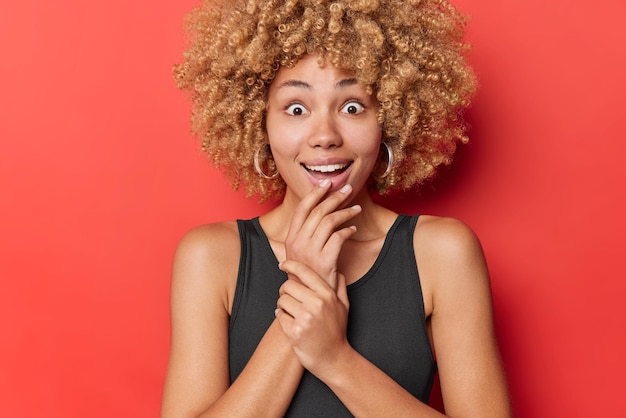 Happy surprised young woman stares with eyes full of interest keeps hand on chin glad and shocked to hear good news wears black t shirt isolated over red background Human reactions concept