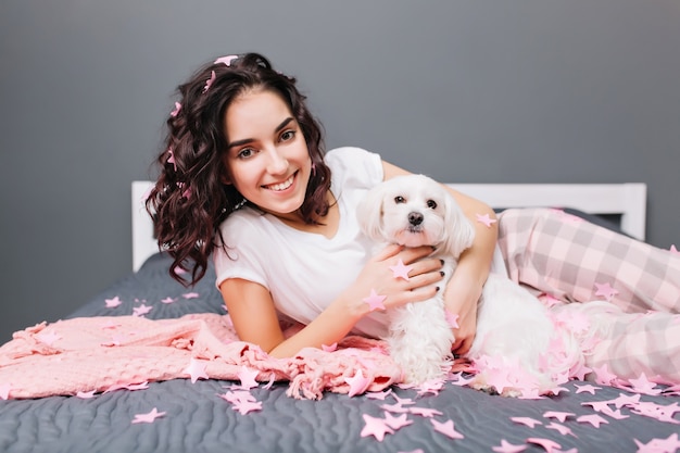 Happy sweet moments of young beautiful woman in pajamas with cut brunette curly hair chilling on bed with little dog  in modern apartment. Smiling in pink tinsels, relaxing at home cosiness