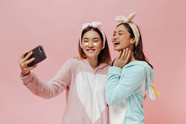 Happy tanned brunette women in pajamas headbands take selfie laugh on pink isolated background