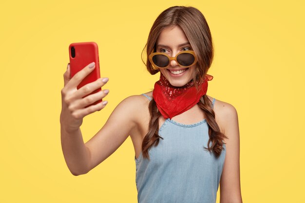 Happy teeanage girl in stylish outfit and sunglasses, holds red cell phone in front, makes selfie portrait, smiles gently, poses against yellow wall. Youth, technology and pastime concept