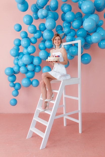 Счастливый подростков девочка, проведение торт ко дню рождения, сидя на белом лестница на розовом фоне