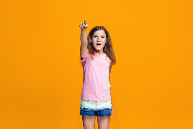 The happy teen girl pointing to you, half length closeup portrait on orange background.