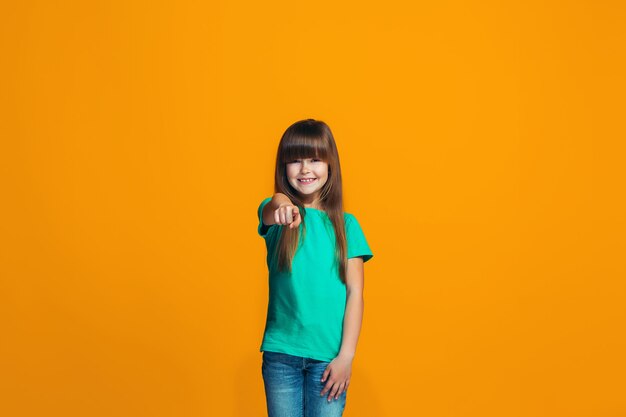 The happy teen girl pointing to you, half length closeup portrait on orange space.