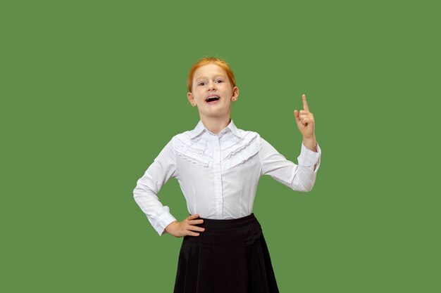 Счастливый подросток девушка стоя, улыбаясь и указывая вверх изолированные на модном зеленом фоне студии. Красивый женский поясной портрет.