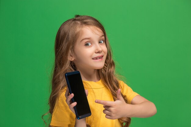 Счастливый подросток девушка стоя, улыбаясь с мобильным телефоном на фоне модных зеленых студии. Красивый женский поясной портрет. Человеческие эмоции, концепция выражения лица.