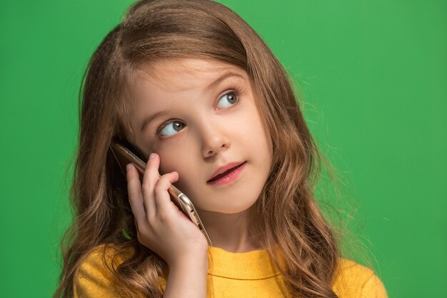 トレンディな緑のスタジオの背景に携帯電話で笑って立って、幸せな十代の少女