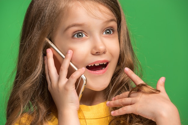 トレンディな緑のスタジオで携帯電話で笑って立っている幸せな十代の少女