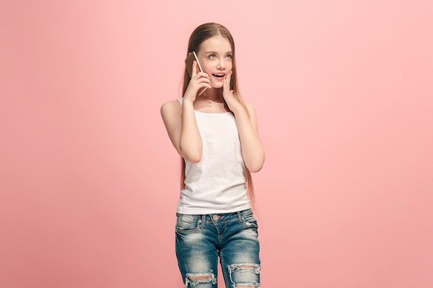 Happy teen girl standing, smiling with mobile phone over trendy pink studio background. Beautiful female half-length portrait. Human emotions, facial expression concept.