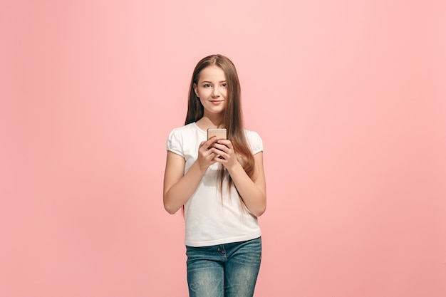 トレンディなピンクのスタジオで携帯電話で笑って立っている幸せな十代の少女