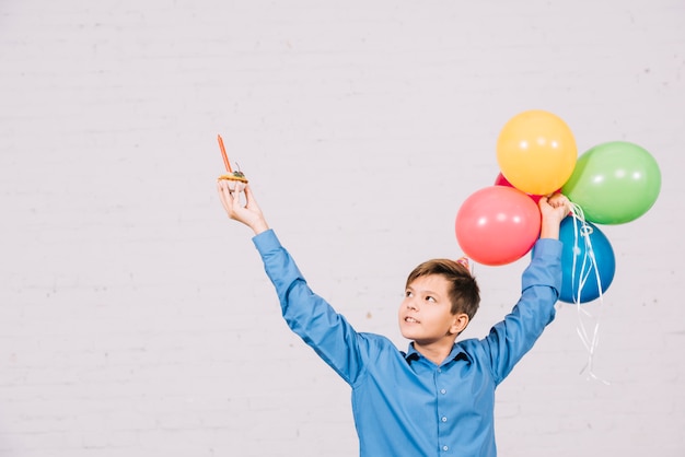 Free photo happy teenage boy holding muffin and colorful balloons raising his hand