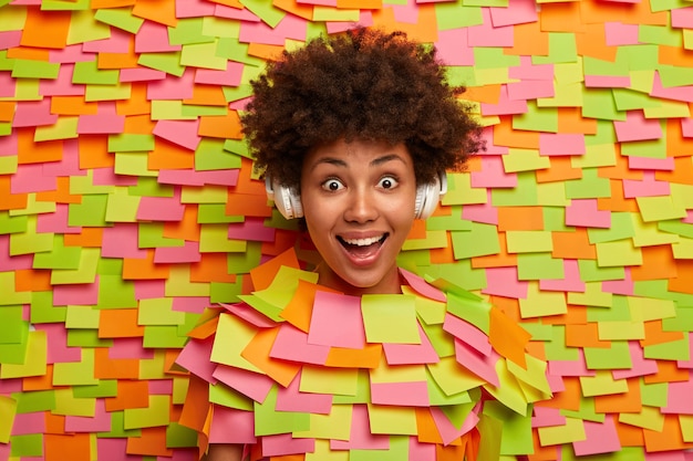 Free photo happy teenage girl has natural curly hair giggles positively listens music in wireless headphones has surprised reaction on stunning news feels upbeat, amused, poses through paper wall adhesive notes