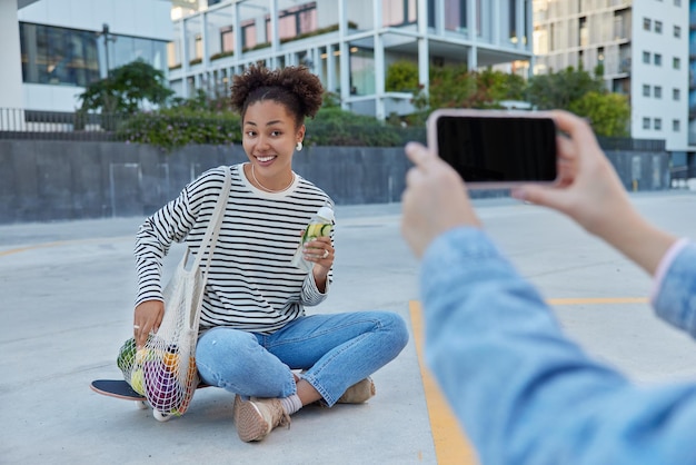 Felice ragazza adolescente pone alla fotocamera dello smartphone per fare foto siede gambe incrociate sullo skateboard porta la borsa a rete con frutta tiene fresca bevanda disintossicante dagli agrumi trascorre il tempo libero in luogo urbano