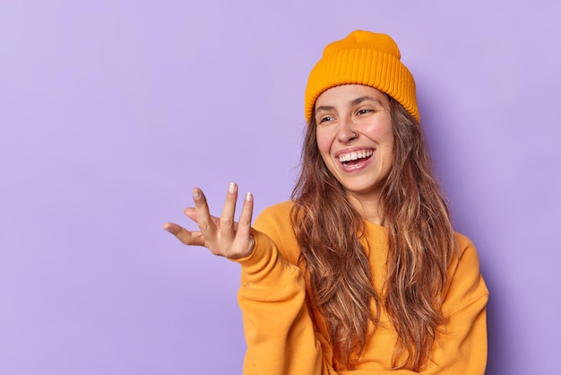 Happy teenage girl raises hand looks joyfully at something funny wears hat and jumper poses against purple background with blank space for your advertising content. Positive human emotions concept