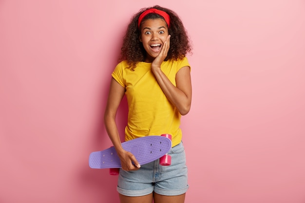 Happy teenage girl with Afro hair holds skateboard, has fun with friend while skating, enjoys active rest