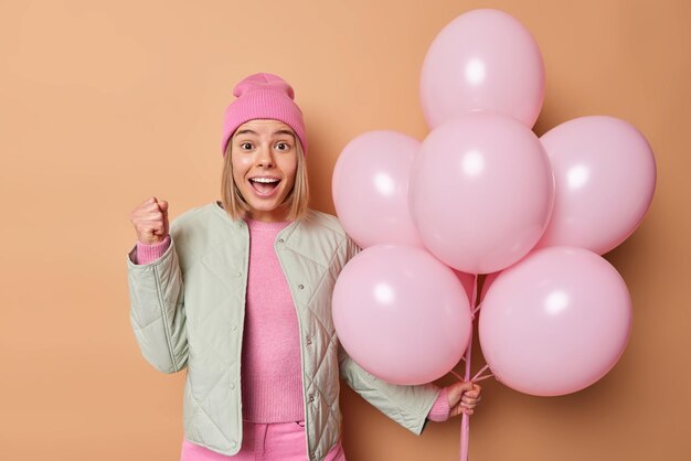 Happy teenage girl with fair hair clenches fist celebrates good news dressed in jacket and pink hat holds bunch of inflated balloons celebrates special occasion isolated over brown background.