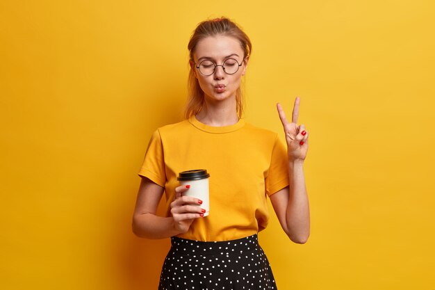 Happy teenager has fun indoor, keeps lips folded, eyes closed, makes peace gesture, enjoys drinking take out coffee, wears bright yellow t shirt, optical glasses, poses indoor