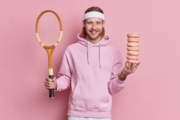 幸せなテニスプレーヤーは健康的なライフスタイルと有害な食べ物のどちらかを選択しますラケットを保持し、甘いドーナツの山はスウェットシャツとヘッドバンドを着用します。バドミントンゲームをするつもりのヨーロッパのひげを生やした男