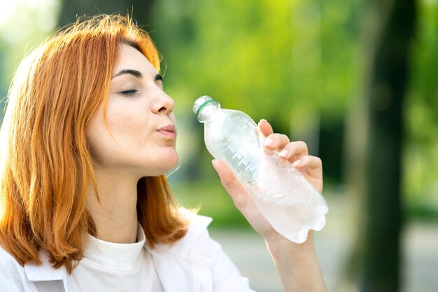 Счастливая жаждущая рыжая женщина пьет свежую воду в бутылках летом на открытом воздухе.