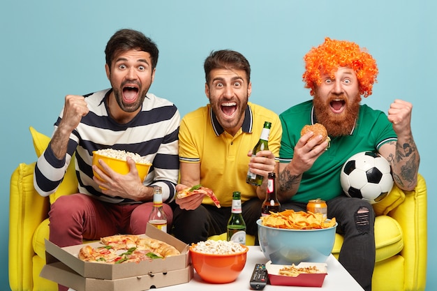 Happy three friends being football fans, watch soccer, pose on sofa in living room, eat fast food, drink cold beer