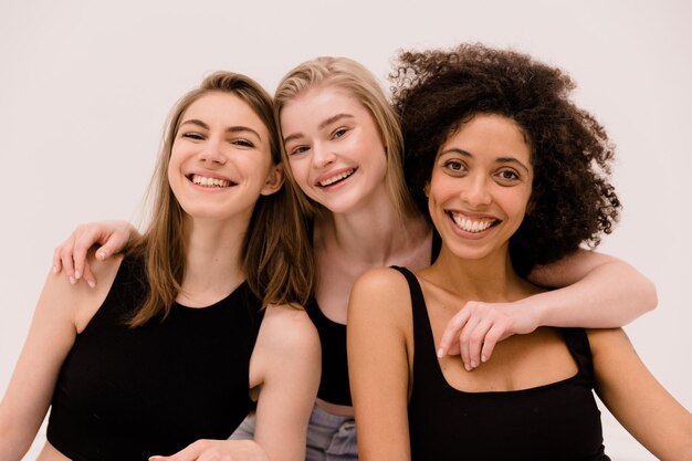 Happy three young caucasian and african women in black tops look at camera on white background