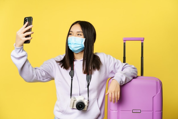 Happy tourist, asian girl on vacation taking selfie in medical mask near suitcase, photographing on smartphone, yellow background