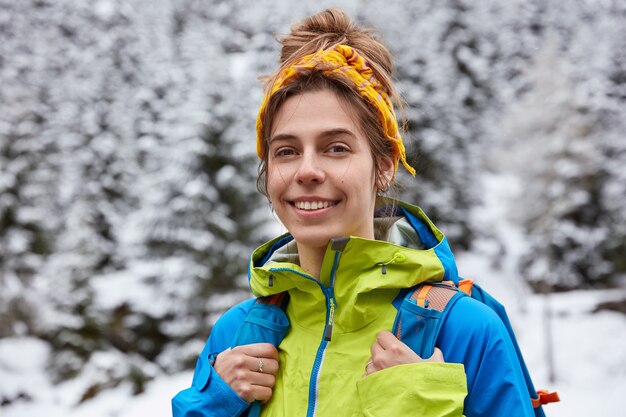 Happy tourist poses on snowy mountain top, enjoys winter day trekking, wears yellow headband, casual jacket