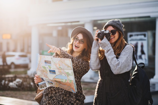 Happy travel together of two fashionable women in sunny city centre. Young joyful women expressing positivity, using map, vacation with bags, camera, making photo, cheerful emotions, great mood.