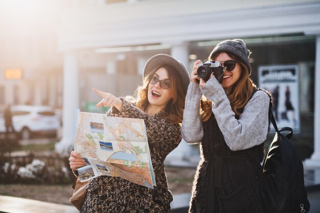 Happy travel together of two fashionable women in sunny city centre. Young joyful women expressing positivity, using map, vacation with bags, camera, making photo, cheerful emotions, great mood.