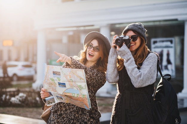 Счастливого путешествия вместе двух модниц в солнечном центре города. Молодые радостные женщины, выражающие позитив, с помощью карты, отдых с сумками, фотоаппарат, фотографирование, веселые эмоции, отличное настроение.
