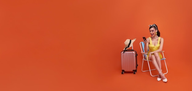 Счастливая туристка-путешественница сидит на стуле с чемоданом с помощью мобильного телефона