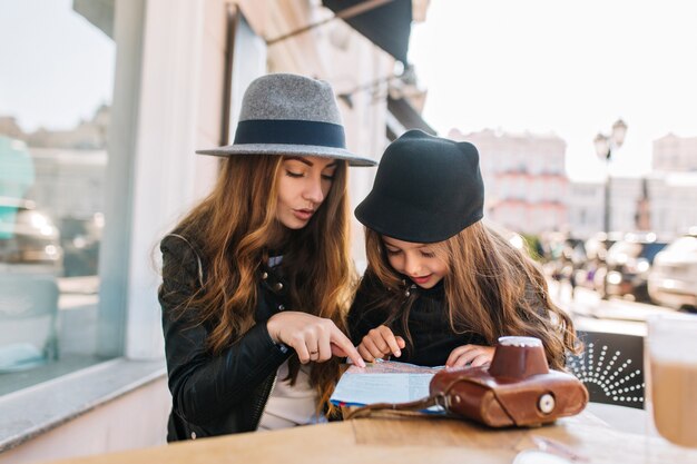 Happy traveling family. Young mother with daughter sitting in cafe on the sunny city background, look at the map. Mom and child having fun at the table.They are stylishly dressed, good mood..