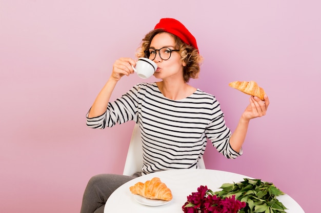 Free photo happy traveling woman in france eating croissans with coffee, sits by the table on pink