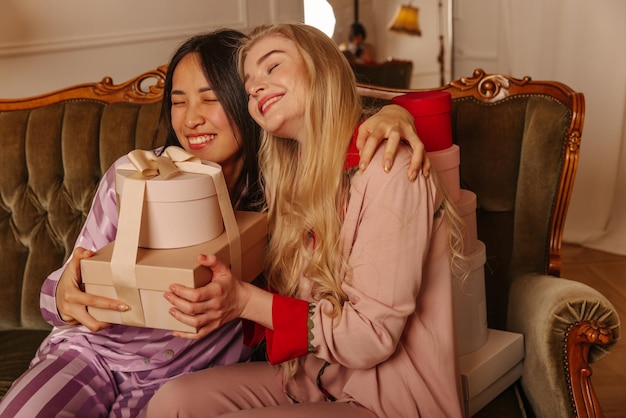 幸せな2人の異人種間の若い女の子がソファに抱き締めてギフトボックスを持って座っている誕生日パーティーで人々の感情の概念
