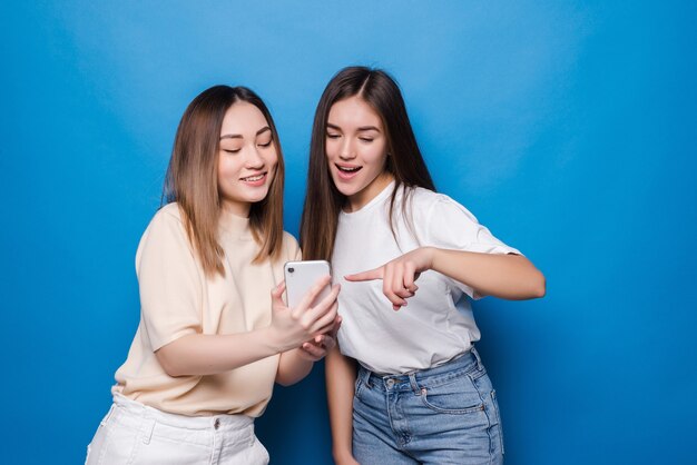 Happy two young girls laughing and pointing finger at screen of smartphone while taking selfie isolated over blue wall