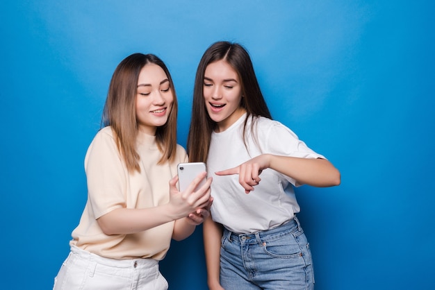 Happy two young girls laughing and pointing finger at screen of smartphone while taking selfie isolated over blue wall