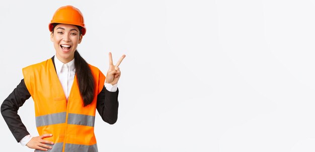 Happy upbeat asian female asian architect in safety helmet and reflective jacket showing peace sign