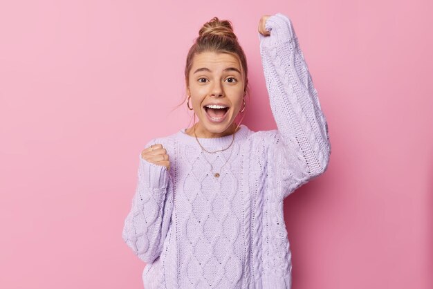 Happy upbeat young European woman with combed hair makes triumph gesture keeps ams raised exclaims yes wears knitted sweater poses against pink background achieves goal or prize Yeah I am winner