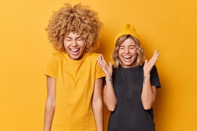 Happy upbeat young female companions laugh out loudly at funny joke feel euphoric express positive emotions dressed casually isolated over vivid yellow background People and happiness concept