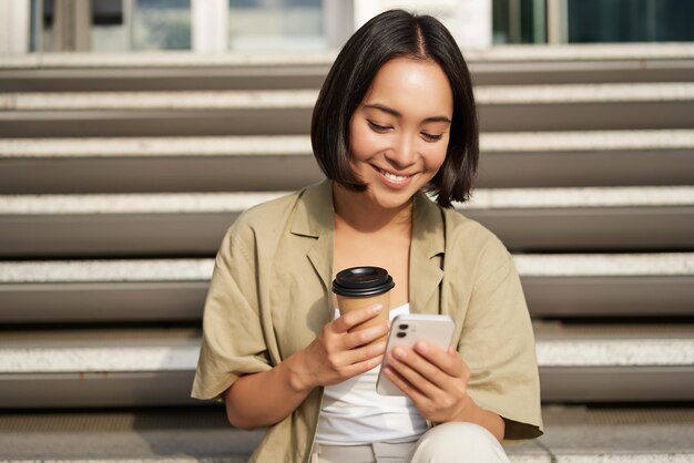 Happy urban girl drinks her takeaway coffee and scrolls feed on smartphone asian woman sits on stair