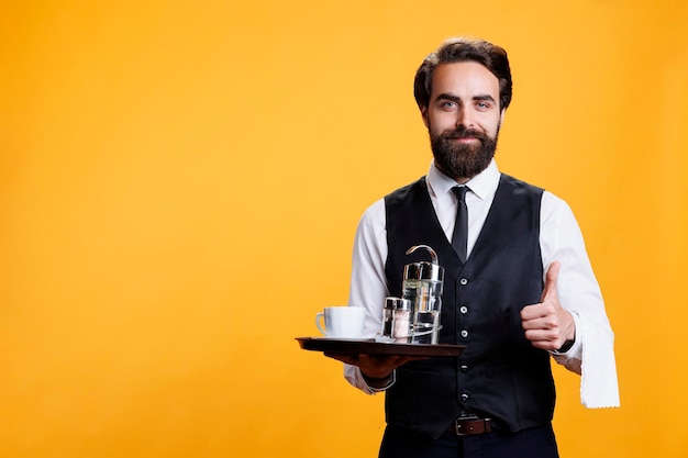 Il cameriere felice fa il segno del pollice in su mentre tiene il piatto con il caffè e gli accessori in studio, il maggiordomo professionale elegante, l'impiegato del ristorante che mostra un gesto di accordo alla telecamera.