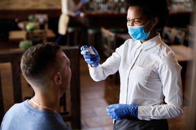 Happy waitress measuring guest's temperature with infrared thermometer in a cafe