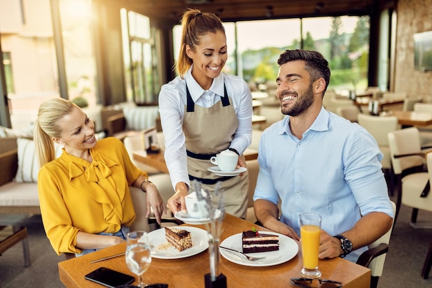 カフェでケーキを食べているカップルにコーヒーを提供する幸せなウェイトレス