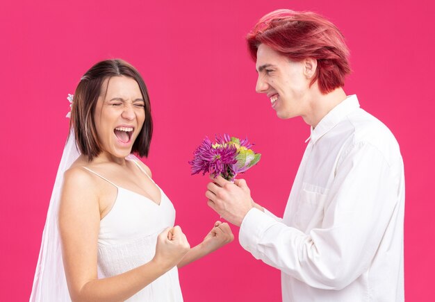 Happy wedding couple groom and bride happy and cheerful groom giving flowers for his excited bride in wedding dress standing on pink