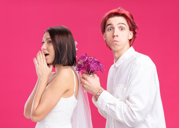 행복한 결혼 커플 신랑과 신부 행복하고 쾌활한 신랑이 핑크색 벽 위에 서 있는 웨딩 드레스를 입고 웃는 신부를 위해 꽃을 주고 있습니다.