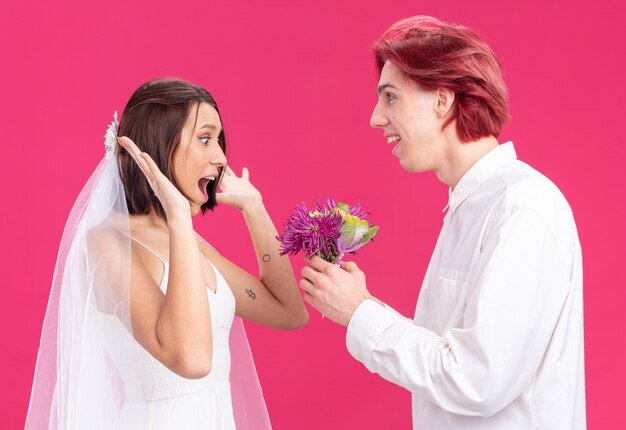 Счастливая свадебная пара жених и невеста счастливый и веселый жених дарит цветы своей улыбающейся и удивленной невесте в свадебном платье, стоящей на розовом
