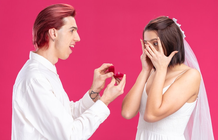 मुफ़्त फोटो happy wedding couple of groom and bride man making propose with wedding ring in a gift box happy and excited standing on pink