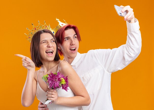 Happy wedding couple groom and bride with bouquet of flowers in wedding dress wearing gold crowns smiling cheerfully doing selfie using smartphone